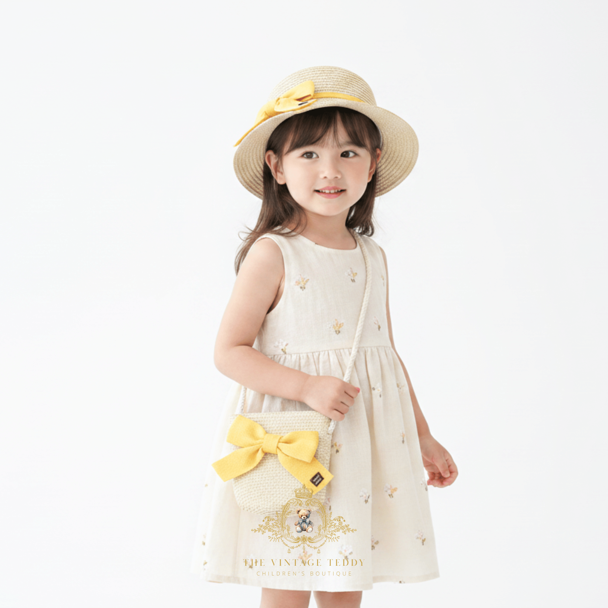 A young girl wearing a straw sun hat and matching handbag, smiling in a cream  children's dress, the kids accessories is adorned with a yellow ribbon, in bright background studio setting. The Vintage Teddy's brand logo is visible at the bottom of the image.