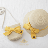 A straw sun hat and matching woven crossbody bag set for children in a light beige tone, both decorated with a large yellow  bow, displayed on a white fabric background.