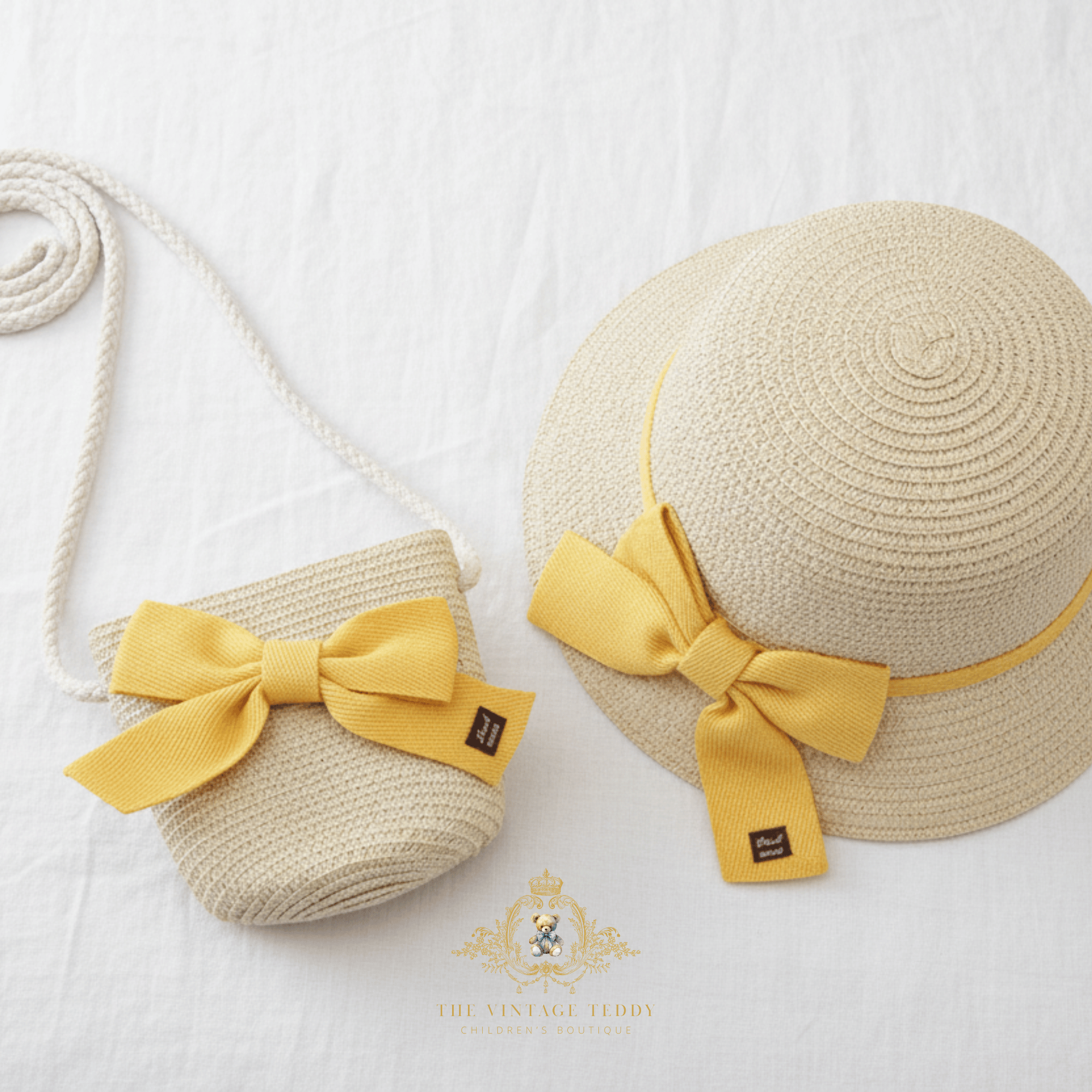 A straw sun hat and matching woven crossbody bag set for children in a light beige tone, both decorated with a large yellow  bow, displayed on a white fabric background.