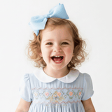 A young girl with curly light blonde hair wearing a baby blue hair bow and baby blue girls smocked dress with a white Peter Pan collar, smiling broadly against a plain white background.