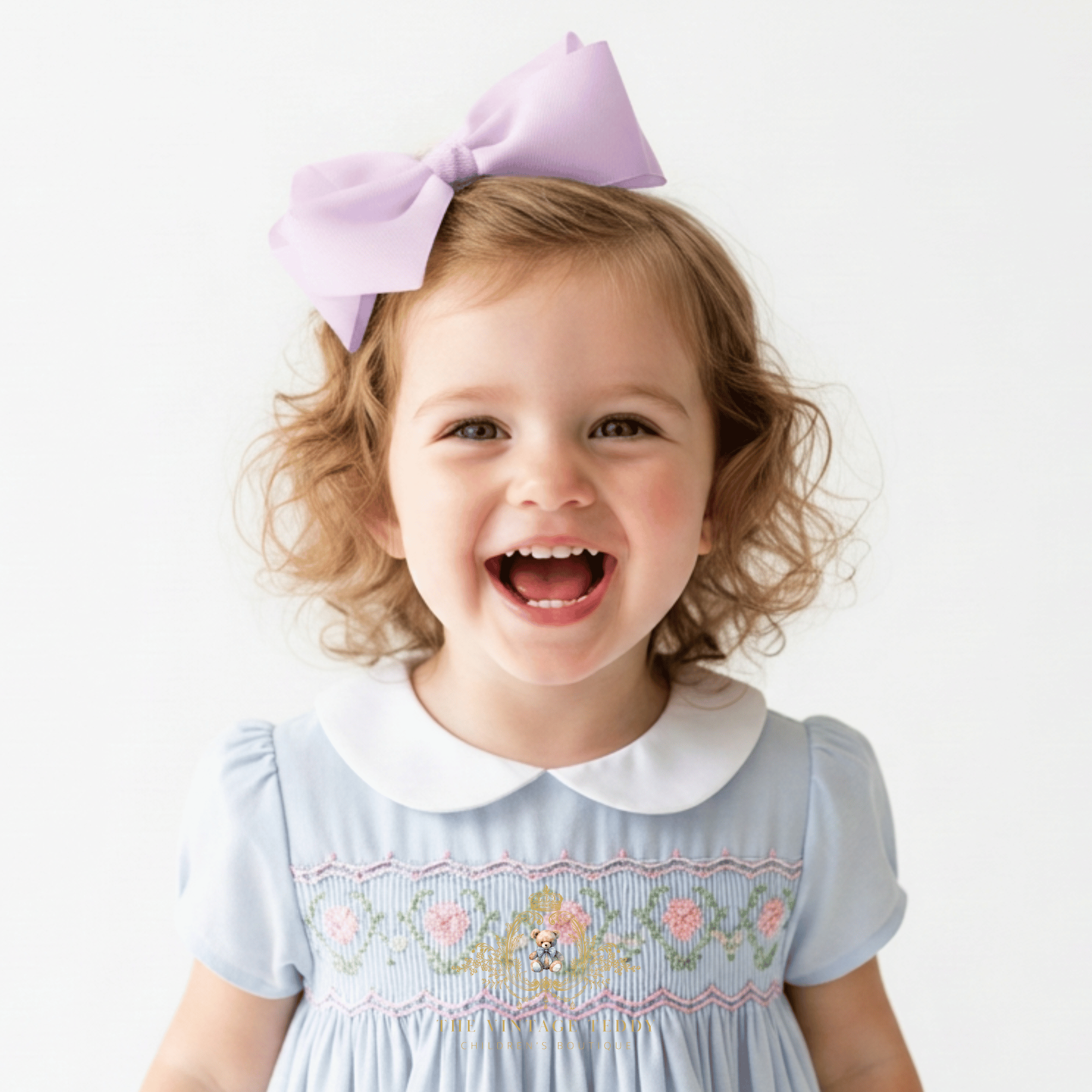 A young child with curly blonde hair wearing a lavender hair bow and a blue little girls smocked dress with a white Peter Pan collar, smiling against a plain white background.