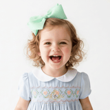 A young girl with curly blonde hair wearing a mint green hair bow and a baby blue kids smocked dress with a white Peter Pan collar, smiling broadly against a plain white background.