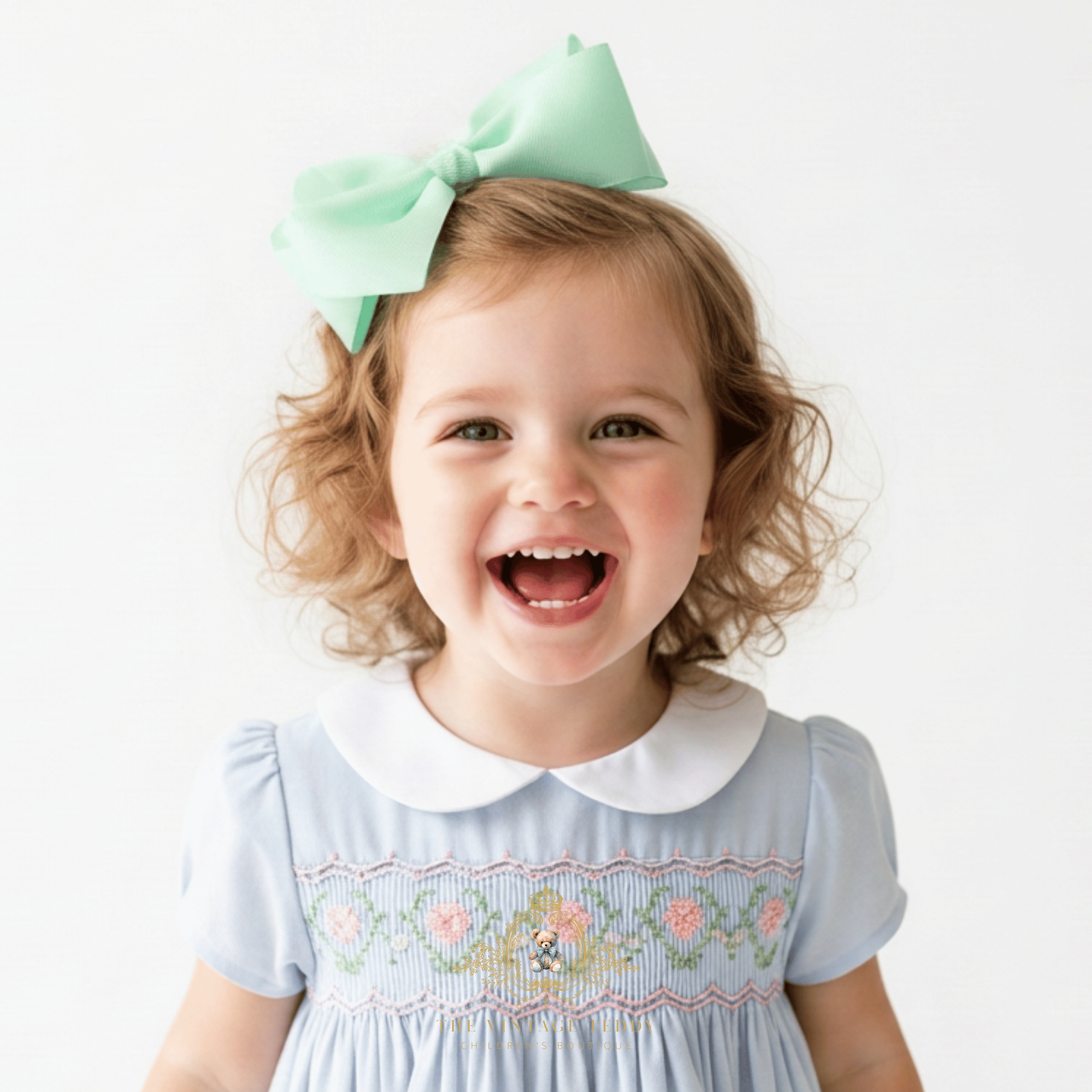A young girl with curly blonde hair wearing a mint green hair bow and a baby blue kids smocked dress with a white Peter Pan collar, smiling broadly against a plain white background.