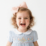A young child with curly blonde hair wearing a light blue girls smocked dress with a white Peter Pan collar and a large pale pink bow, smiling against a plain white background.