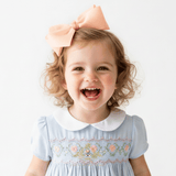 A young girl with curly blonde hair wearing a large peach hair bow and a baby blue smocked dress for children with a white Peter Pan collar, smiling broadly against a plain white background.