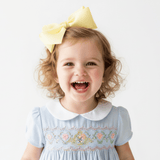 A young girl with curly blonde hair wearing a large pastel yellow hair bow in her hair and a light blue children's embroidered smock dress with a white Peter Pan collar, smiling broadly against a plain white background.