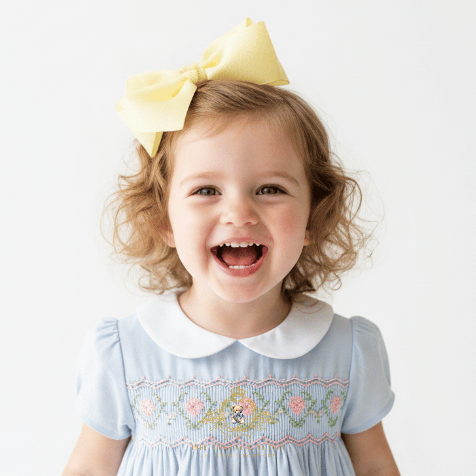 A young girl with curly blonde hair wearing a large pastel yellow hair bow in her hair and a light blue children's embroidered smock dress with a white Peter Pan collar, smiling broadly against a plain white background.