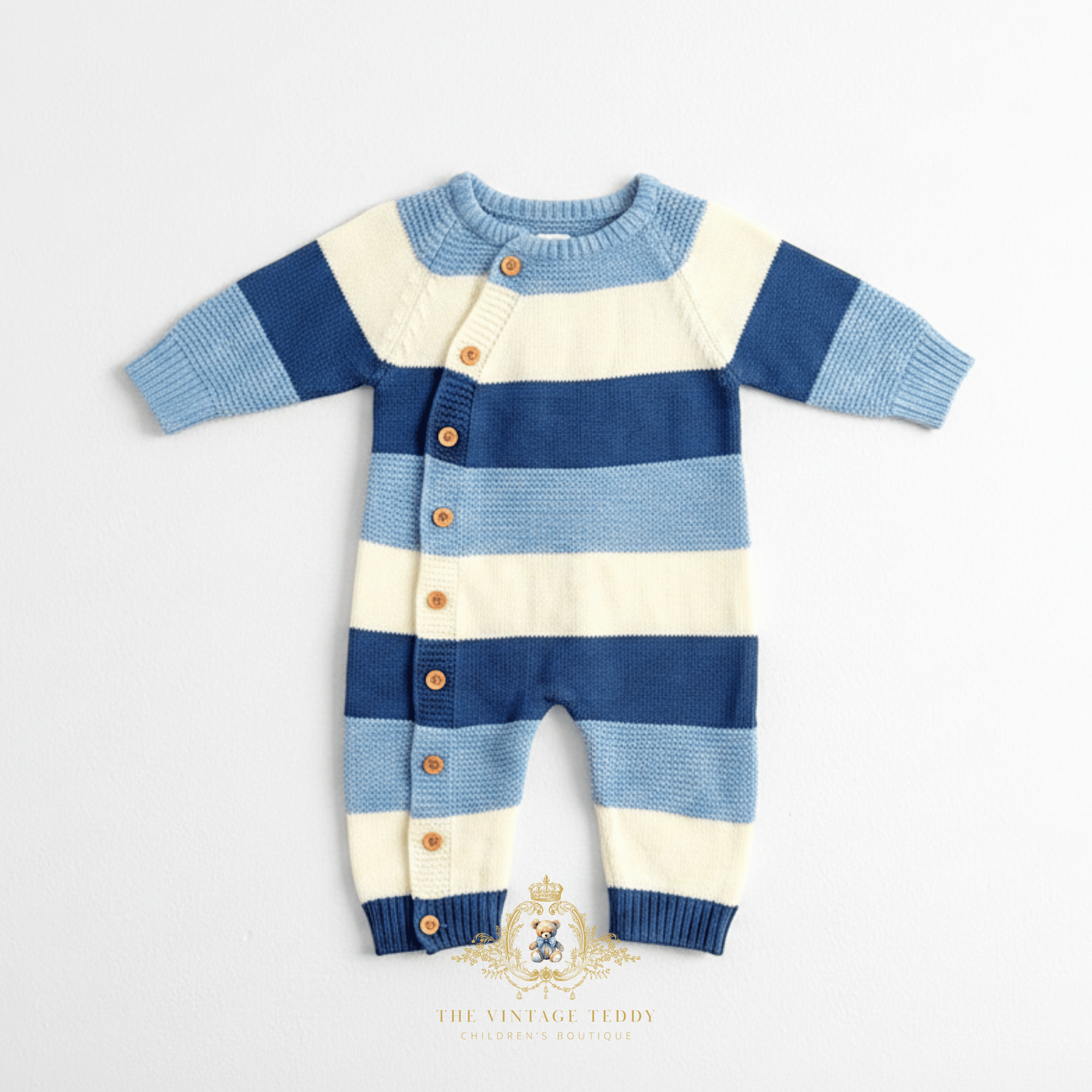 A baby boy knit one-piece romper with horizontal stripes blue and white, featuring a button-down on the side front with wooden buttons and ribbed cuffs and round collar, laid flat on a white background surface. 'The Vintage Teddy watermark logo displayed at the bottom center. 