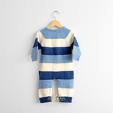 A baby boys knit jumpsuit with horizontal stripes in two-tones of blue and white, hanging on a natural wooden hanger against a plain white wall.