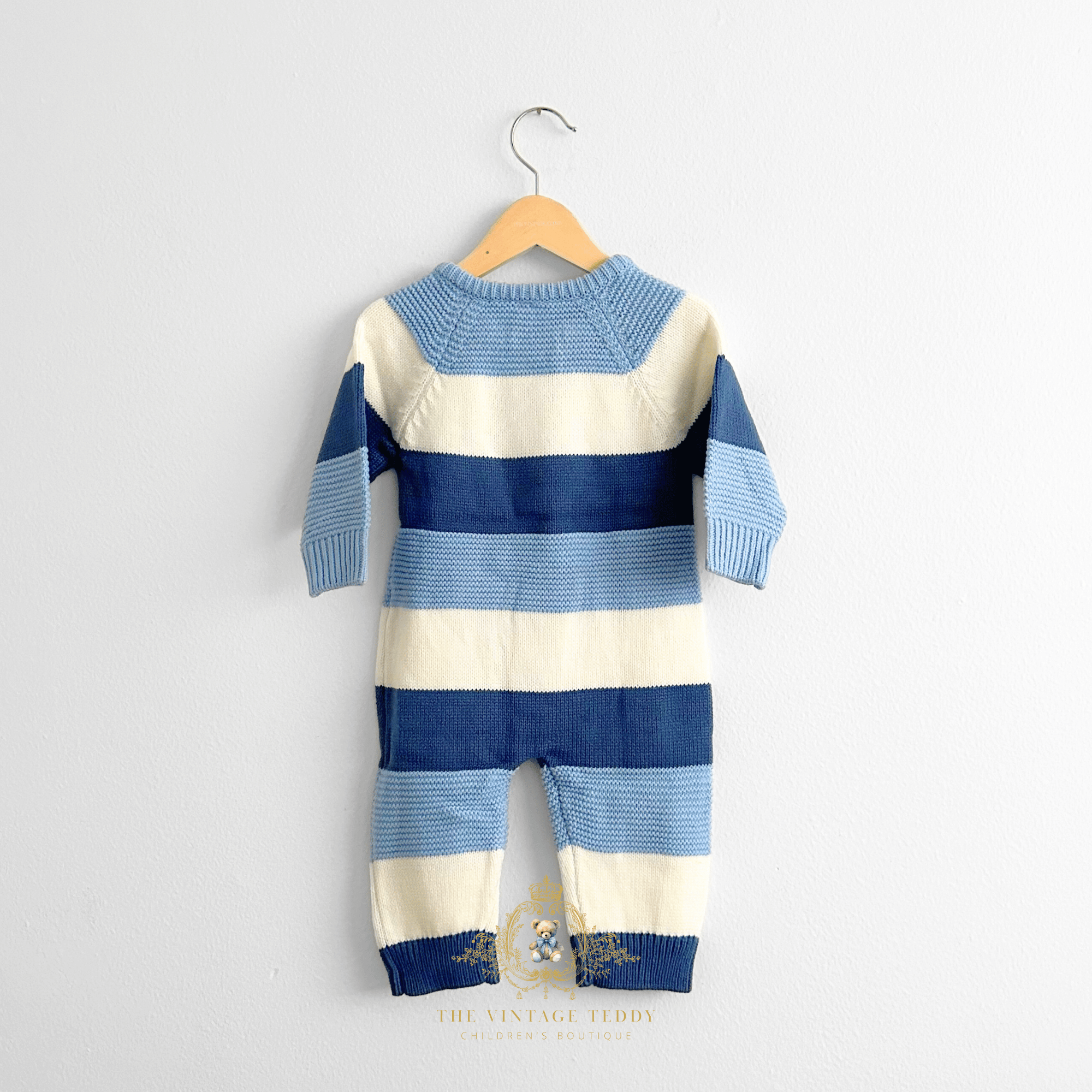A baby boys knit jumpsuit with horizontal stripes in two-tones of blue and white, hanging on a natural wooden hanger against a plain white wall.