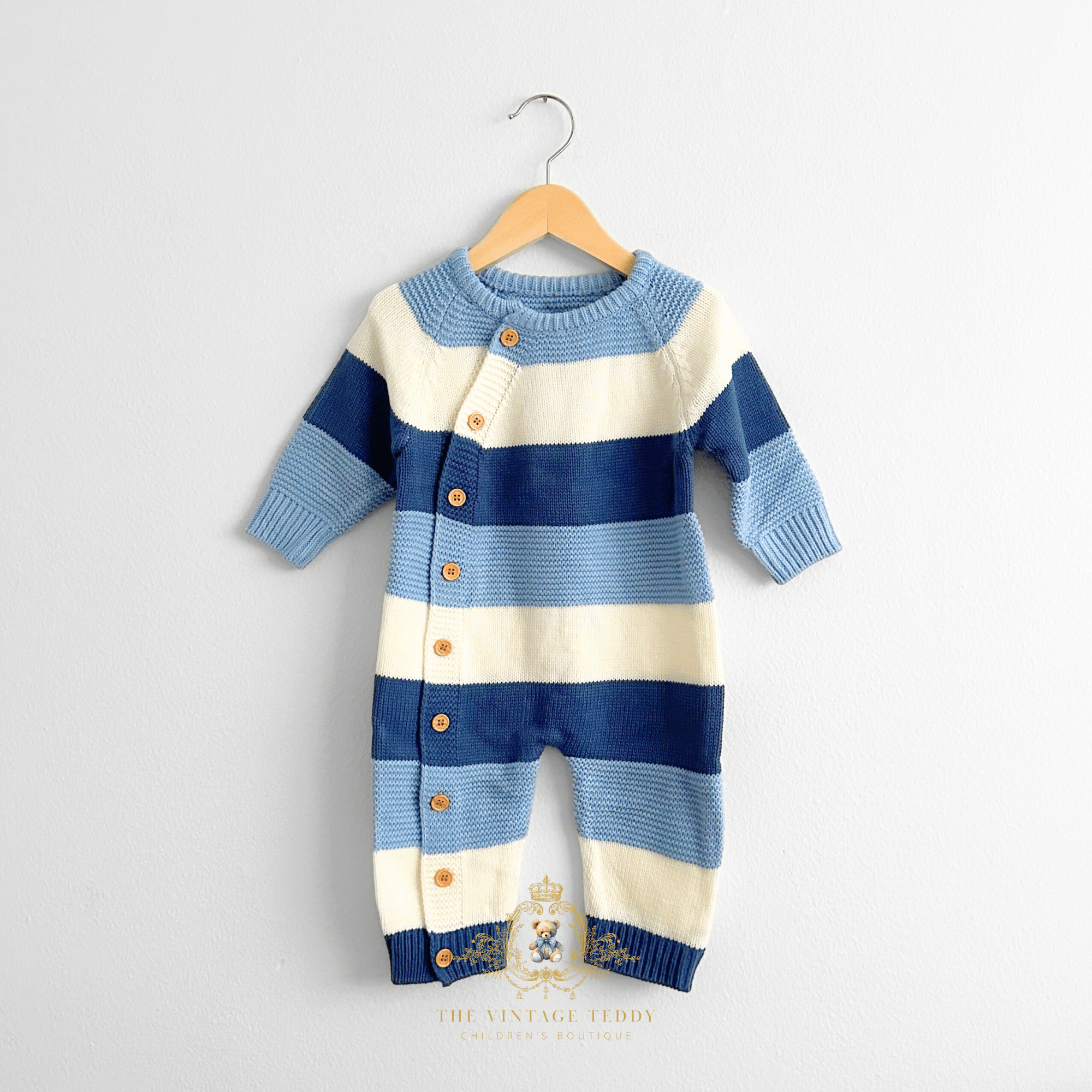 A baby boy knit one-piece romper with horizontal stripes in blues and white, hanging on a natural wooden hanger against a white wall. The baby romper features a button closure down the front side and ribbed cuffs and hem. The Vintage Teddy watermark displayed bottom center.