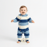 A baby boy standing and smiling while wearing a knitted one-piece romper with horizontal cornflower blue, sky blue, and white stripes. The romper has brown wooden buttons down the front side. Clean white studio background and 'The Vintage Teddy' watermark logo bottom centered.