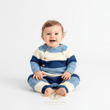 A smiling baby boy sitting wearing a blue and white striped knit one-piece bodysuit,  against a clean white studio background. A watermark at the bottom reads The Vintage Teddy Clothing Boutique.