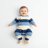 A baby boy lying on a white studio background wearing a knitted long-sleeve romper with horizontal stripes in cornflower blue, sky blue, and white. The romper has brown buttons down the front side and 'The Vintage Teddy' logo watermark is displayed at the bottom of the image.
