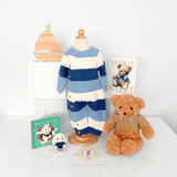 A baby boys knit one-piece outfit displayed on a natural wood mannequin torso, styled with a teddy bear, a small plush toy rattle, a natural wood sailboat, children's books, ABC white wood blocks, and a 'Teddy Sale' sign on a white tabletop surface against a white wall.