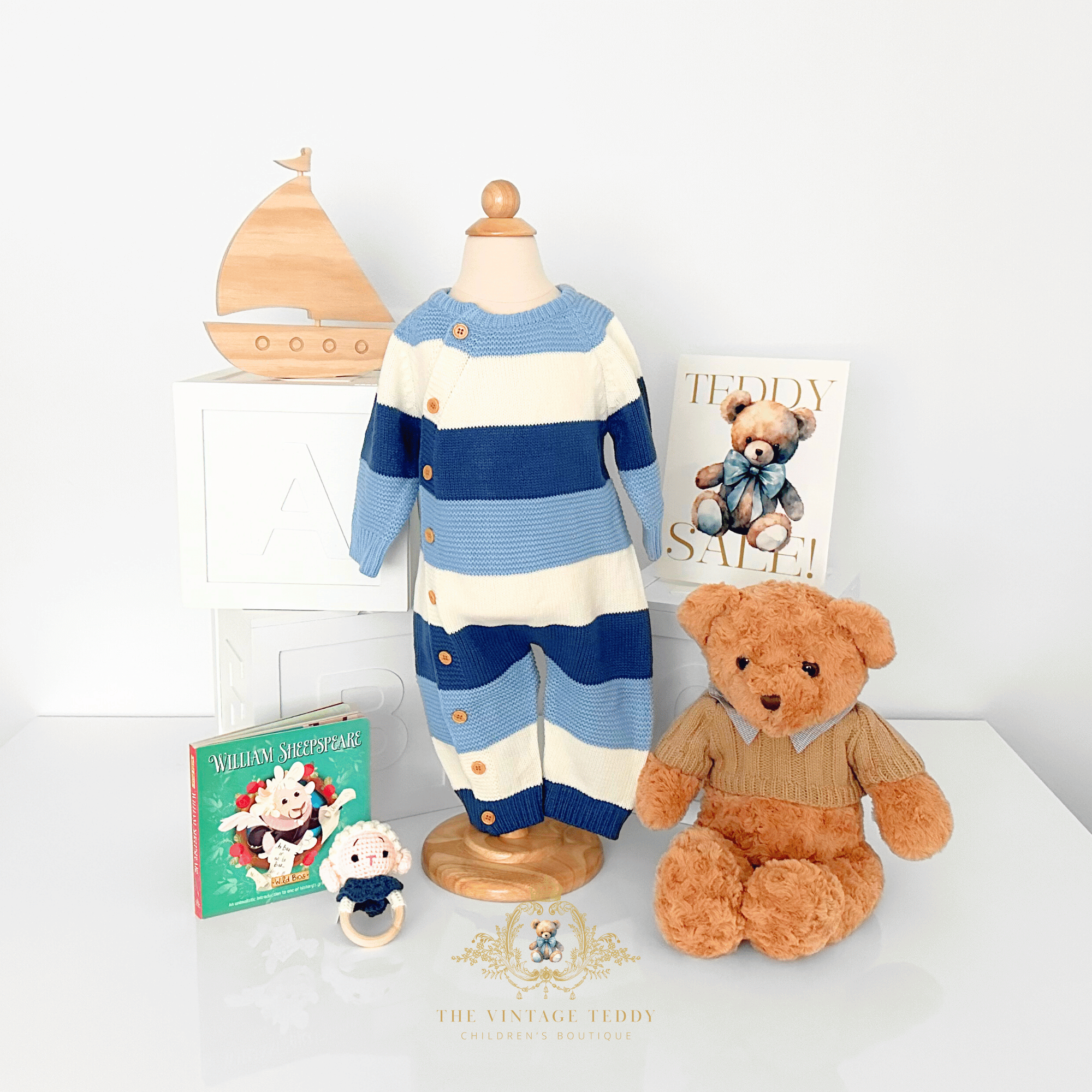 A baby boys knit one-piece outfit displayed on a natural wood mannequin torso, styled with a teddy bear, a small plush toy rattle, a natural wood sailboat, children's books, ABC white wood blocks, and a 'Teddy Sale' sign on a white tabletop surface against a white wall.