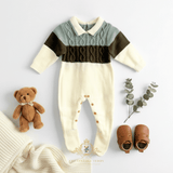A baby boys long-sleeve footed romper laid flat on a white background, styled with a brown teddy bear, eucalyptus sprig, tan baby shoes, and a cream knit blanket. The romper has a cream base with a pale sage and a olive green cable-knit chest panel, and a cream collar.