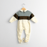 A baby boys long-sleeve footed romper hanging on a natural wooden hanger against a white wall. The baby romper has a sage upper chest and shoulder area, olive green horizontal bands, and cream-colored lower body and sleeve ends.