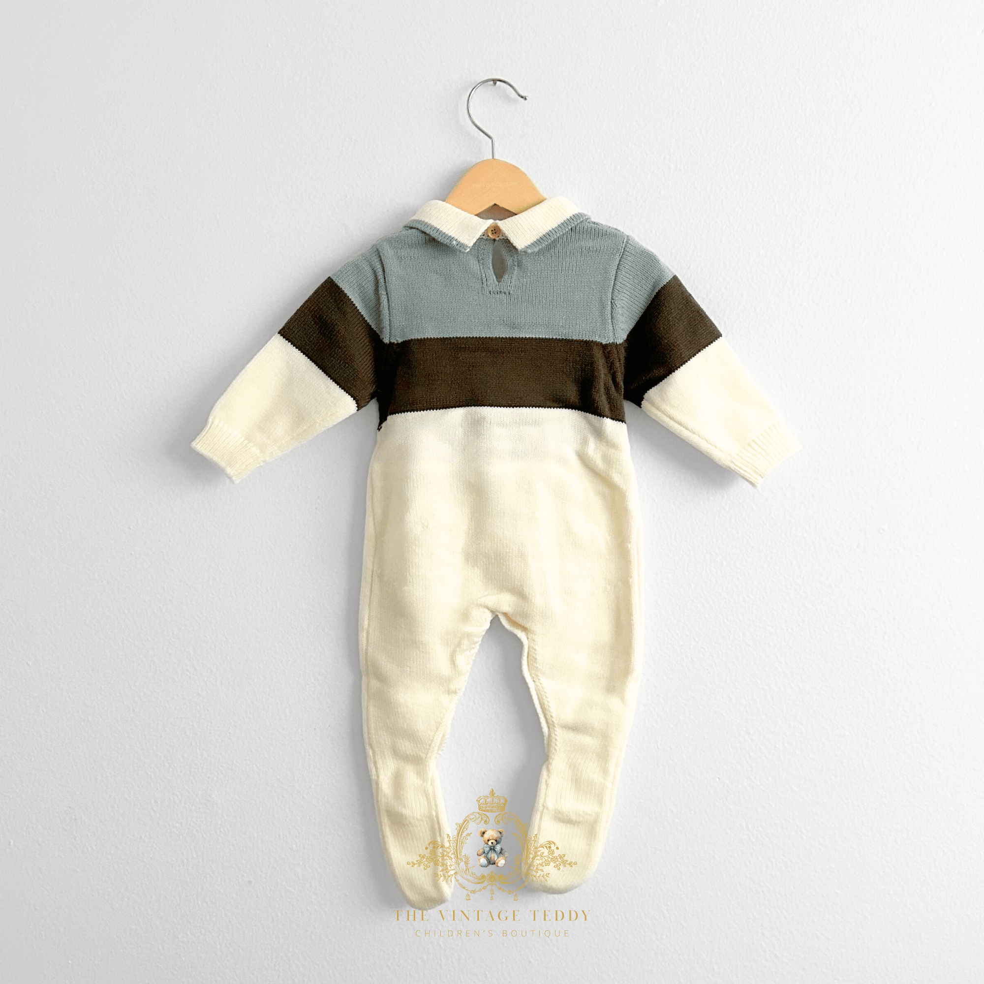 A baby boys long-sleeve footed romper hanging on a natural wooden hanger against a white wall. The baby romper has a sage upper chest and shoulder area, olive green horizontal bands, and cream-colored lower body and sleeve ends.