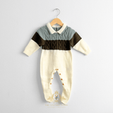 A baby boy knit footed jumpsuit hanging on a natural wooden hanger against a light white wall. The baby jumpsuit has a cream-colored body and legs, a sage and olive green chest panel, and a central cable-knit band.