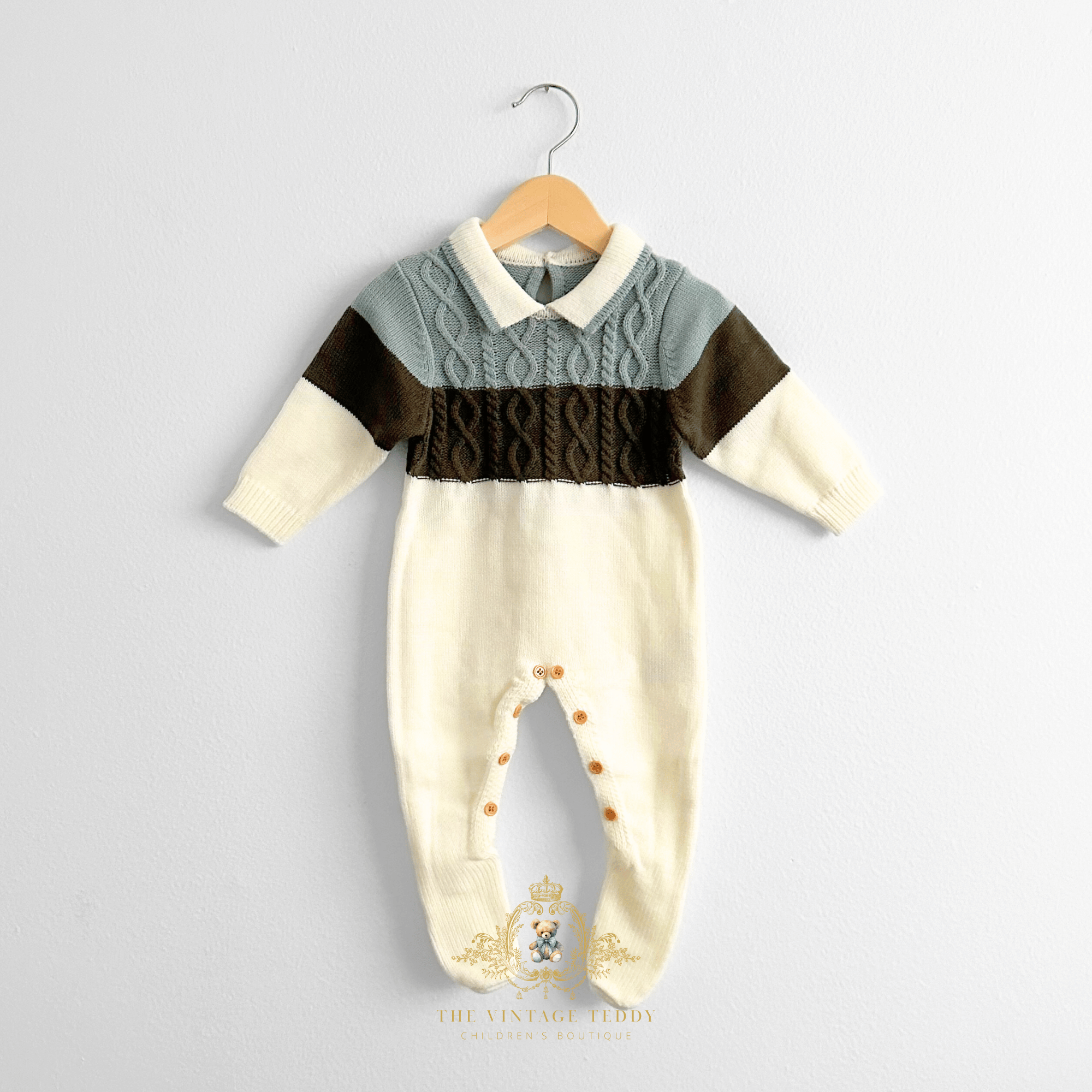 A baby boy knit footed jumpsuit hanging on a natural wooden hanger against a light white wall. The baby jumpsuit has a cream-colored body and legs, a sage and olive green chest panel, and a central cable-knit band.