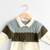 A baby boy long-sleeve knit sweater suit with a color-blocked design, featuring a light sage upper chest, olive green middle band, and cream lower body and sleeves. It has a cable-knit pattern across the chest and is shown hanging on a natural wooden hanger against a white wall.