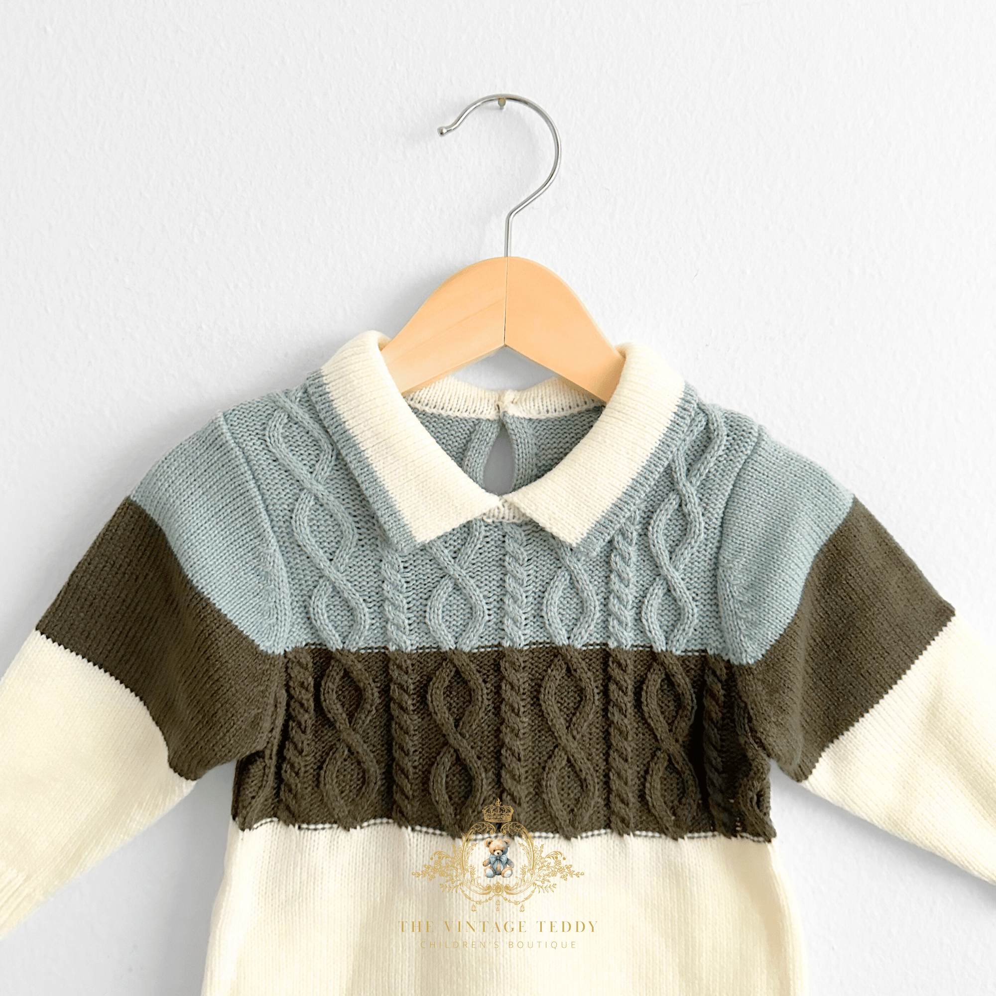 A baby boy long-sleeve knit sweater suit with a color-blocked design, featuring a light sage upper chest, olive green middle band, and cream lower body and sleeves. It has a cable-knit pattern across the chest and is shown hanging on a natural wooden hanger against a white wall.