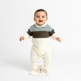 A smiling baby boy standing and wearing a cream knit footed one-piece with a sweater-style romper in sage and olive green. The one-piece has button closures on the legs and a small decorative brand  logo at the bottom.
