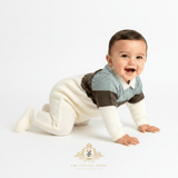 A baby crawling on a white studio floor wearing a cream, sage and olive green  knit footed one-piece. The brand logo 'The Vintage Teddy Clothing Boutique' is visible at the bottom.