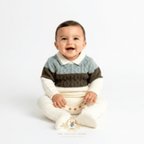 A smiling baby boy sitting, wearing a knit collared sweater one-piece footie baby romper in sage, olive green, and cream. The photo is taken against a seamless white studio background with a watermark 'The Vintage Teddy' logo at the bottom.