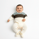 A smiling baby boy lying on their back wearing a cream-colored knit footed bodysuit with a color-blocked chest design in sage, olive green and cream. The image is shot against a clean white background with a watermark 'The Vintage Teddy' logo at the bottom.