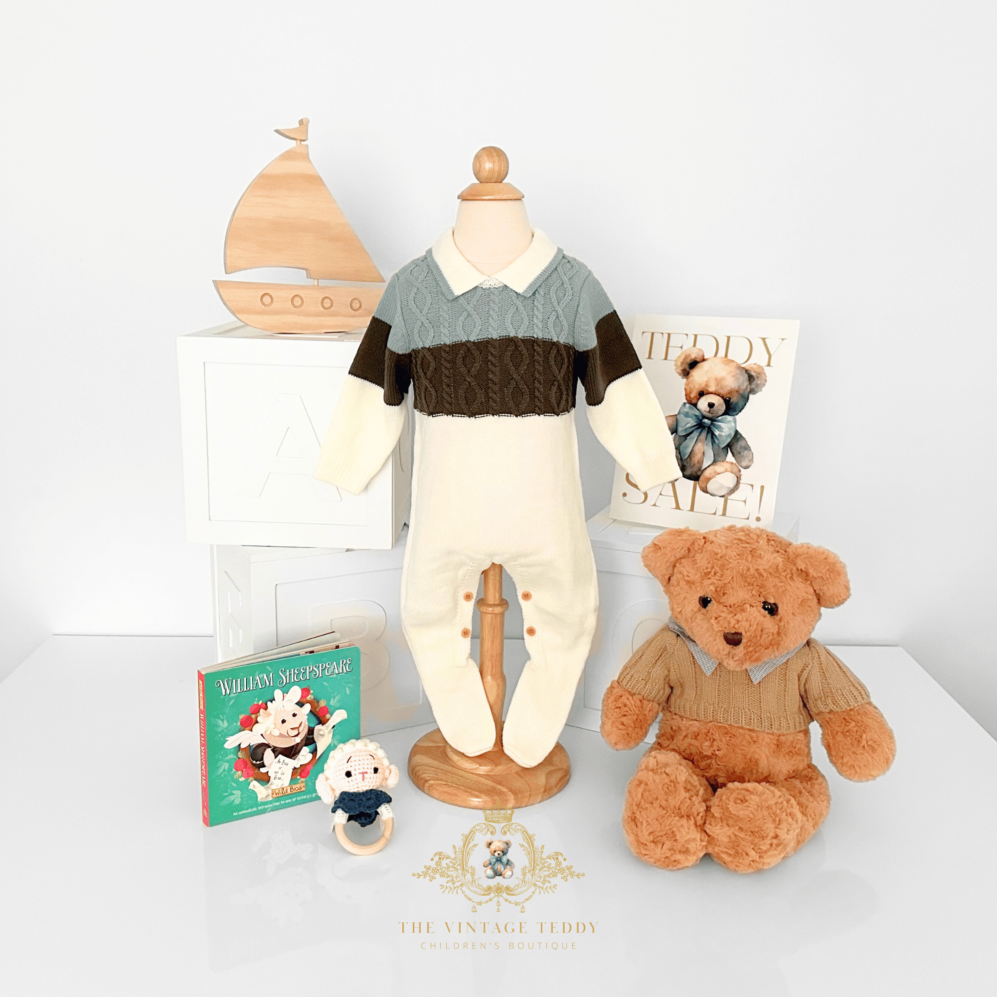 A baby footed knit romper displayed on a wooden mannequin, styled with a teddy bear, a small plush toy rattle, a children's book, a natural wood sailboat, white ABC wooden blocks decoration and a 'Teddy Sale' sign against a clean white background.