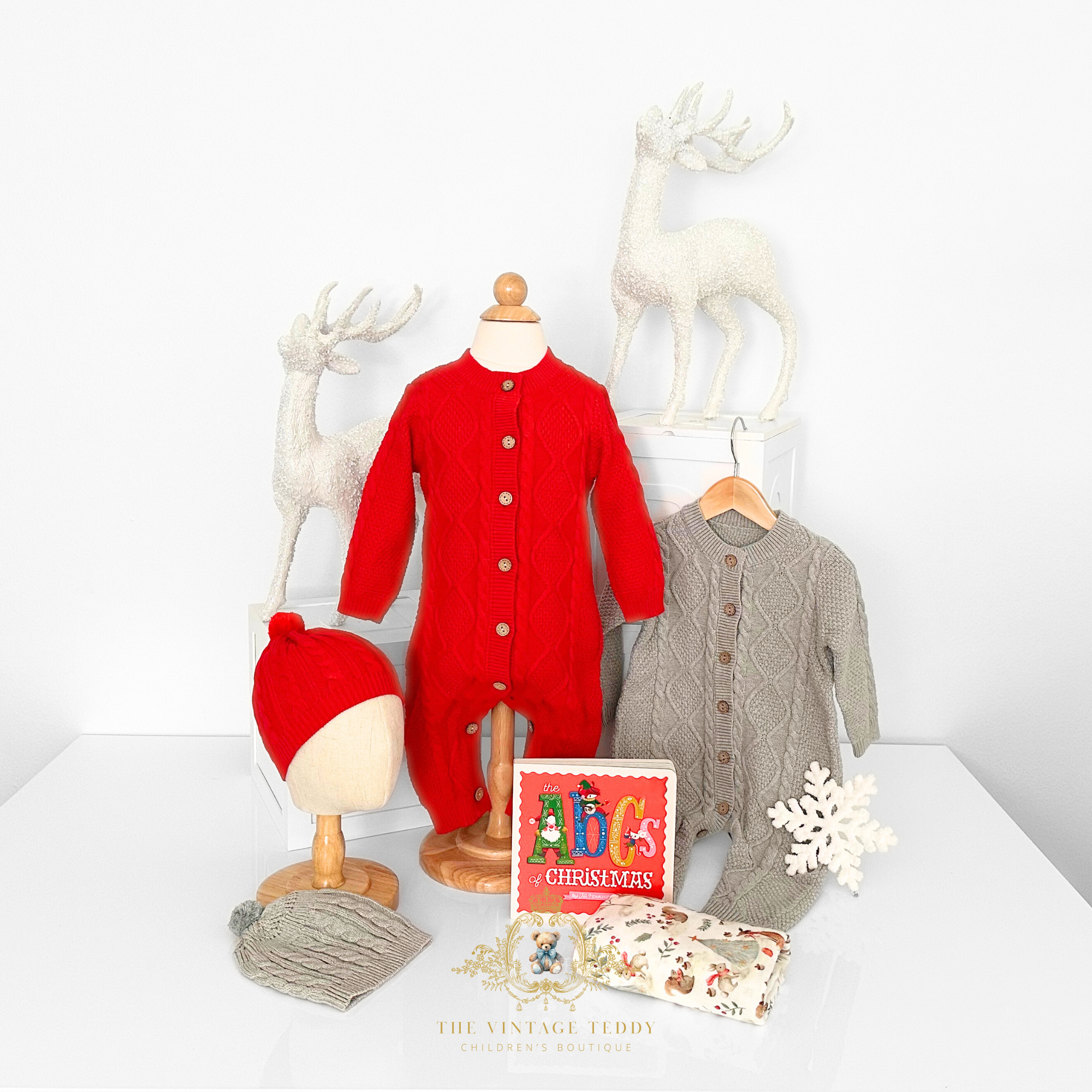 Children's red and gray cable knit one-piece bodysuit, matching caps, children's book, and blanket, decorative Christmas items on a white tabletop display with white deer statues on wooden ABC blocks.