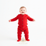 Baby girl wearing a bright red cable-knit button-front romper, standing on a seamless white studio background. The brand logo 'The Vintage Teddy Clothing Boutique' is visible at the bottom.