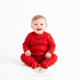 Smiling baby girl sitting on a white studio background, wearing a bright red cable-knit one-piece with front buttons. 'The Vintage Teddy' boutique decorative watermark /logo appears near the bottom center.