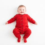 Smiling baby girl lying on her back wearing a bright red cable-knit romper with button closures, photographed against a clean white background.