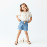 A young girl stands against a white studio background wearing a cream puff-sleeve top with blue floral embroidery and blue denim shorts, along with light brown sandals and a blue hair bow. A watermark logo for The Vintage Teddy Clothing Store is visible at the bottom.