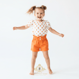 A young girl with her hair in two buns jumps with arms outstretched, wearing a white short-sleeve top with orange pumpkin prints and bright orange shorts, against a plain white background.
