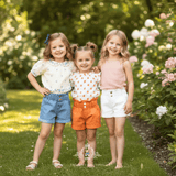 Three young girls standing together on a grassy garden path, wearing coordinated summer outfits for kids. All three girls are wearing matching high-waisted denim shorts in blue, white and orange. Logo watermark at the bottom that reads The Vintage Teddy.