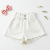 Girls white high-waisted shorts for kids with high waistband and two silver buttons, styled in a flat lay with a pink bow, pearl hair clips, and The Vintage Teddy's brand watermark at the bottom.