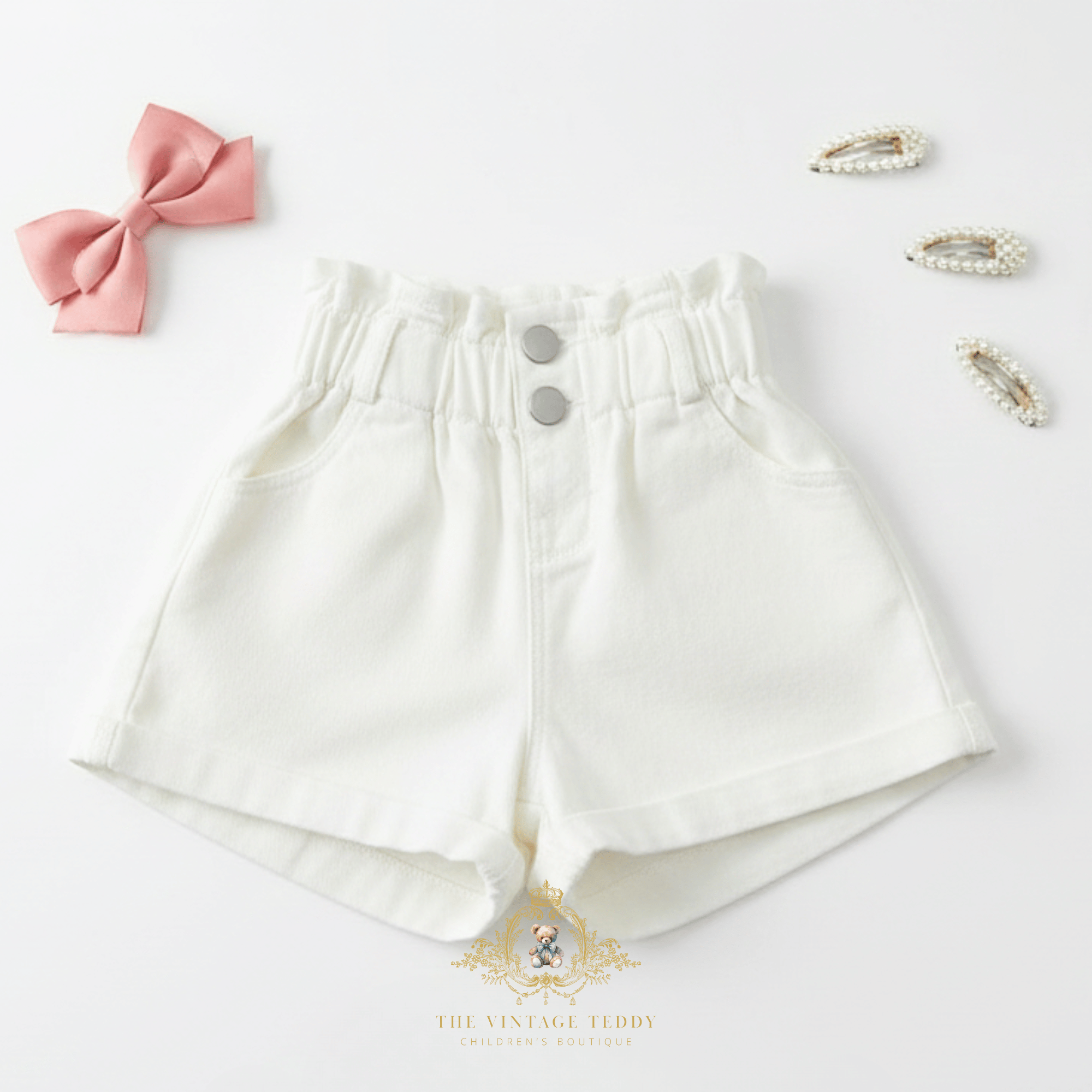 Girls white high-waisted shorts for kids with high waistband and two silver buttons, styled in a flat lay with a pink bow, pearl hair clips, and The Vintage Teddy's brand watermark at the bottom.