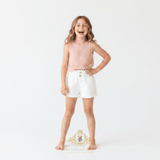 A young girl laughing while wearing a sleeveless light pink knit top and white kids shorts, standing against a plain white studio background. A decorative 'The Vintage Teddy' watermark is visible at the bottom.
