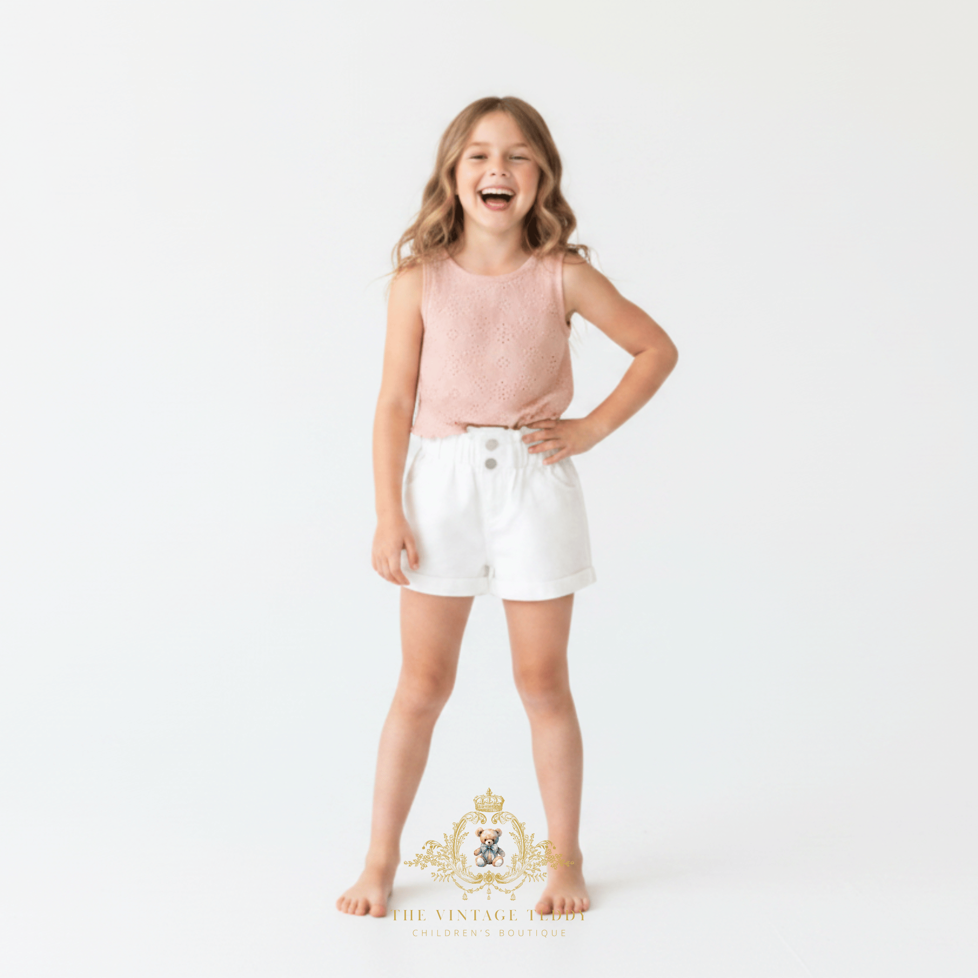 A young girl laughing while wearing a sleeveless light pink knit top and white kids shorts, standing against a plain white studio background. A decorative 'The Vintage Teddy' watermark is visible at the bottom.