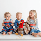 Three happy children sitting together on a white surface against a white background, with a large stuffed turkey toy in the center. The child on the left wears a blue baby romper with a turkey motif, the center child wears a blue long-sleeve shirt with a turkey embroidery, and the child on the right wears a blue-and-white striped long-sleeve girls dress with a blue ruffled skirt.