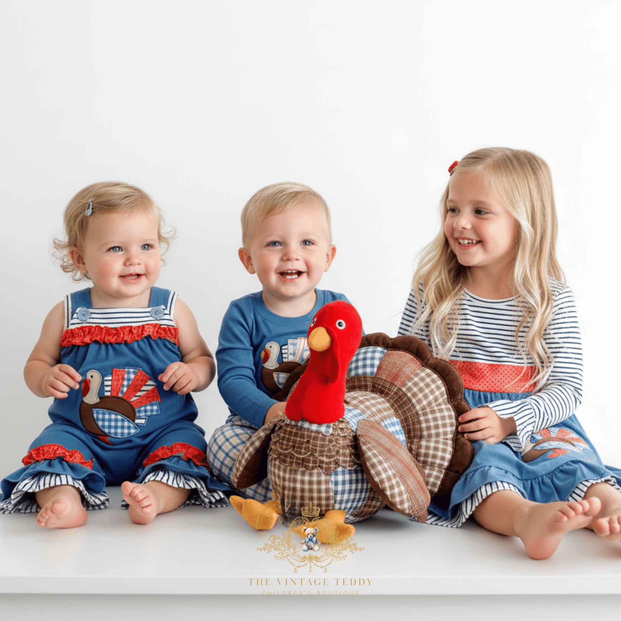Three happy children sitting together on a white surface against a white background, with a large stuffed turkey toy in the center. The child on the left wears a blue baby romper with a turkey motif, the center child wears a blue long-sleeve shirt with a turkey embroidery, and the child on the right wears a blue-and-white striped long-sleeve girls dress with a blue ruffled skirt.