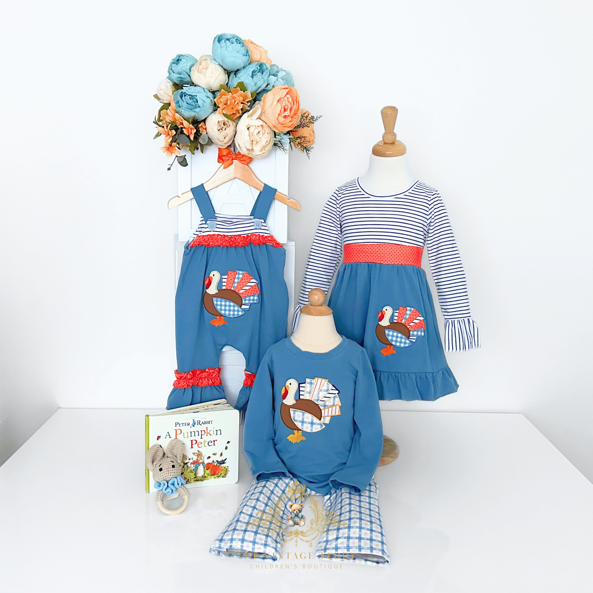A coordinated children's clothing set displayed on a mannequin against a white wall and tabletop decorated with peony flower bouquet, children's book and toy. The set includes a blue sleeveless baby romper with a turkey graphic and orange ruffle, a blue-and-white striped long-sleeve girls dress, and boys matching blue shirt and blue plaid pants set.