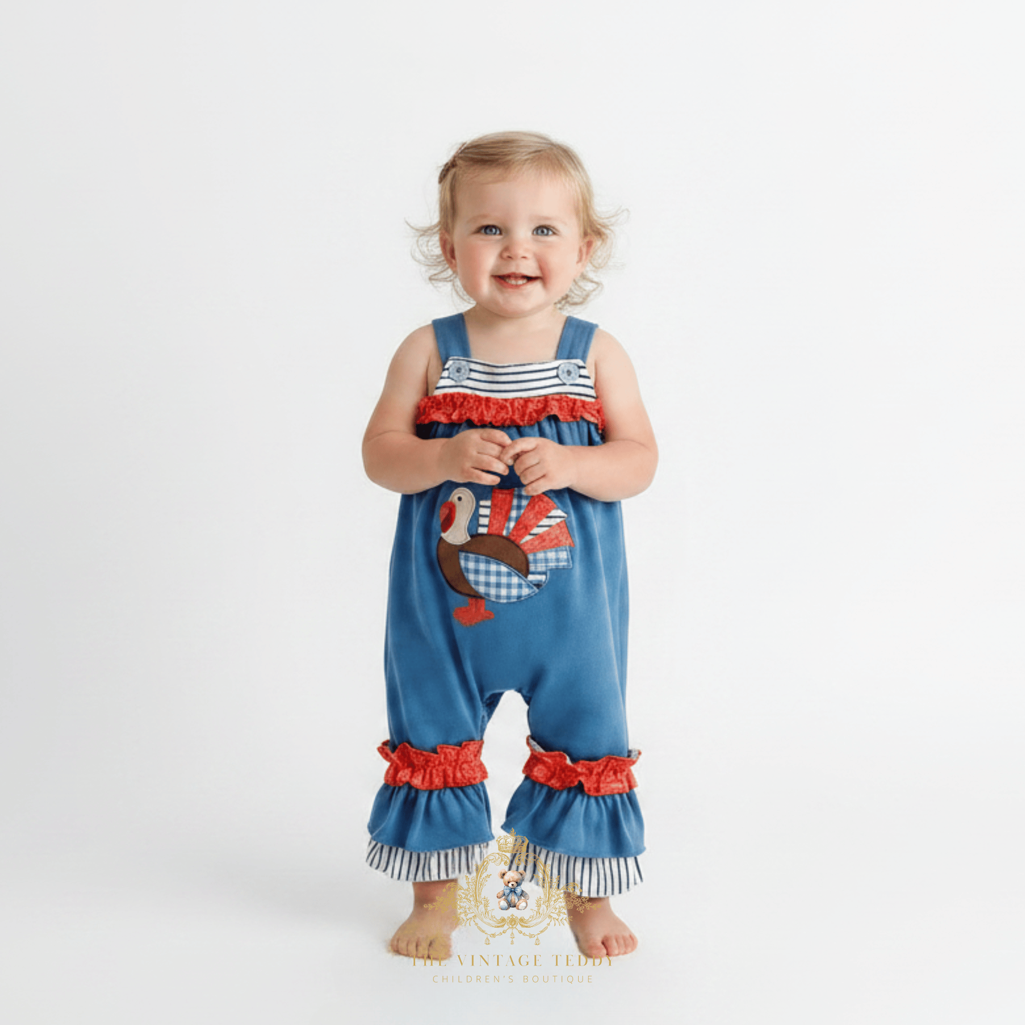 A toddler baby girl stands barefoot against a plain white studio background wearing a blue one-piece romper with orange  ruffles and stripes, shoulder bands, and a patchwork embroidered turkey applique on the front.