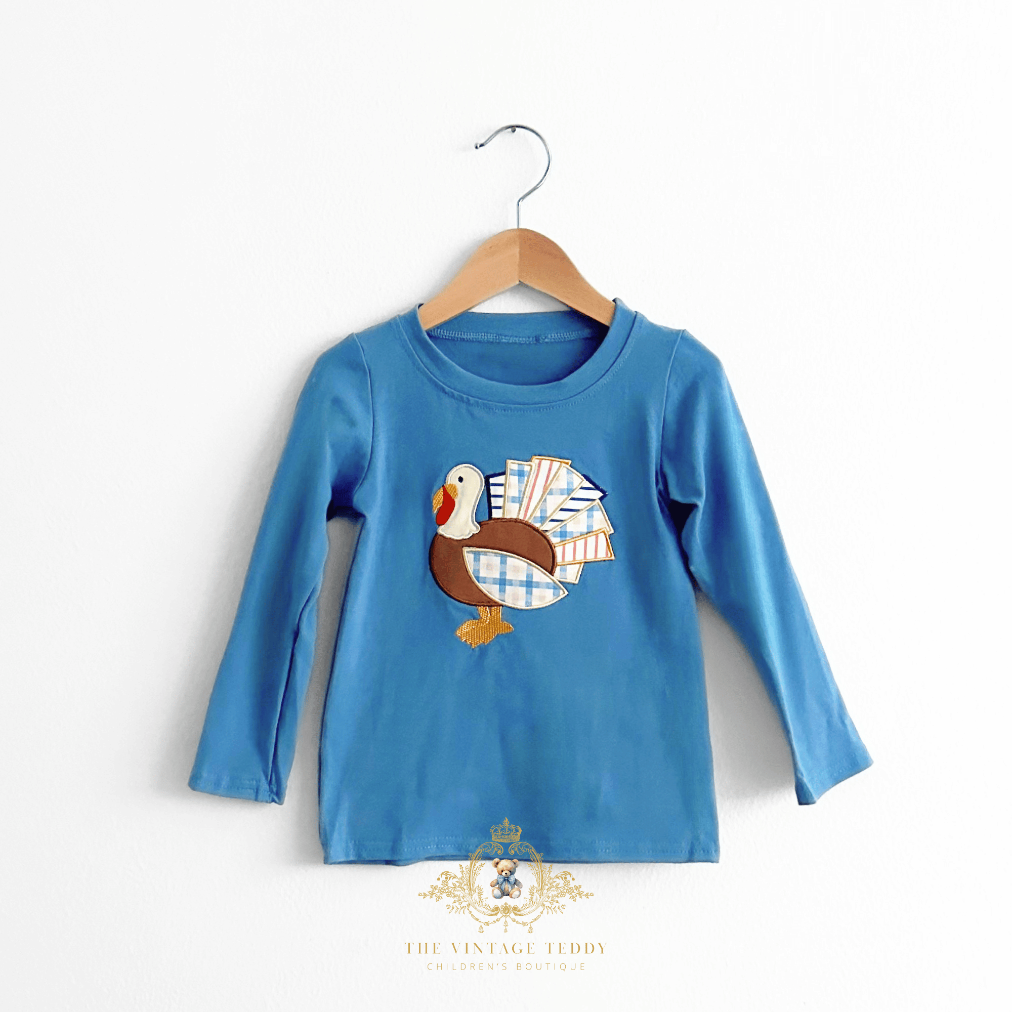 A blue long-sleeve children's shirt hanging on a wooden hanger against a white wall. The boys shirt features a turkey applique embroidery on the chest with a plaid and striped patchwork tail.