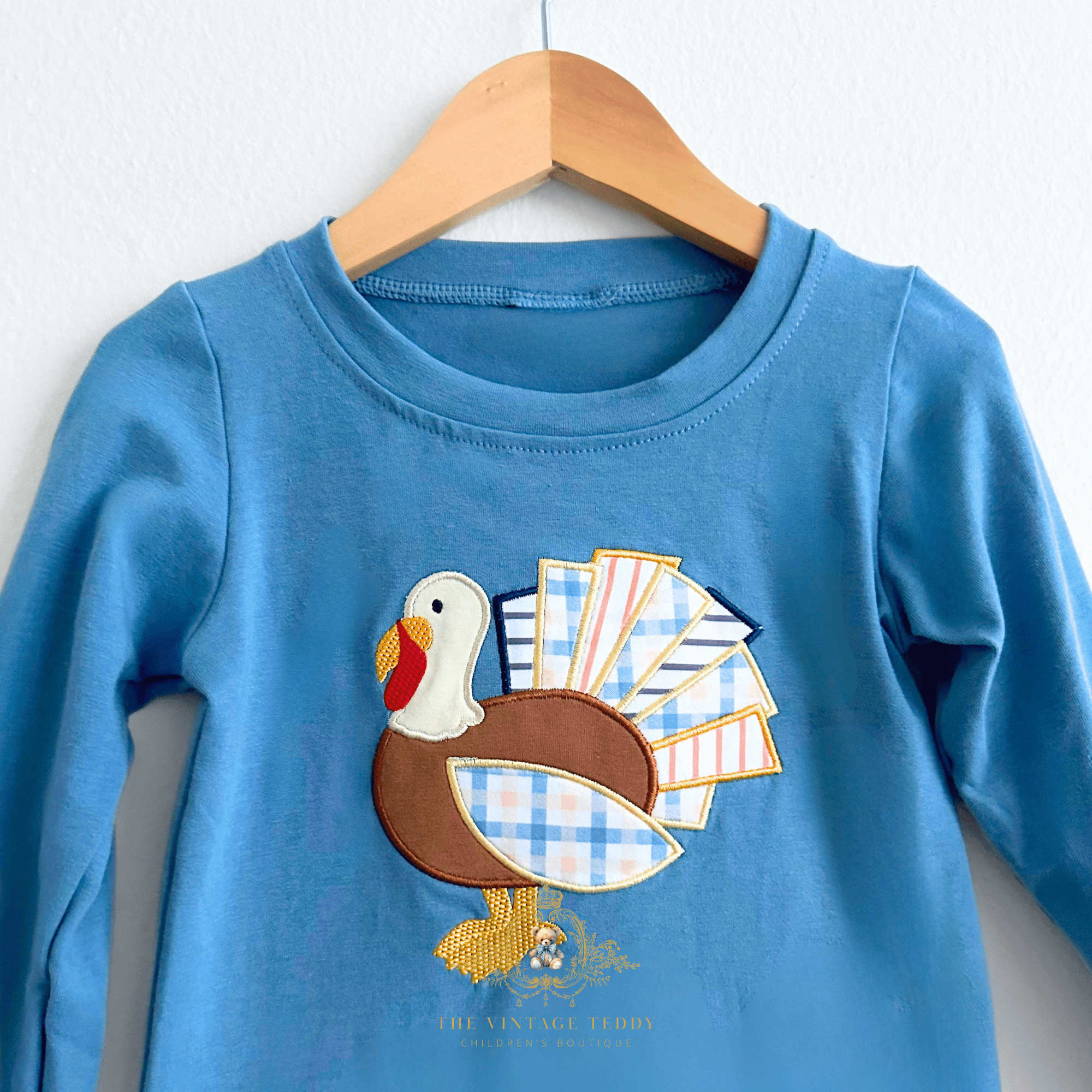 A toddler's long-sleeve blue shirt for boys hanging on a wooden hanger against a white wall, featuring a colorful applique turkey embroidery design on the front. Gobble, Gobble!