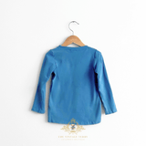 The back of a kids blue long-sleeve top hanging on a wooden hanger against a plain white background, with a watermark at the bottom reading 'THE VINTAGE TEDDY'.